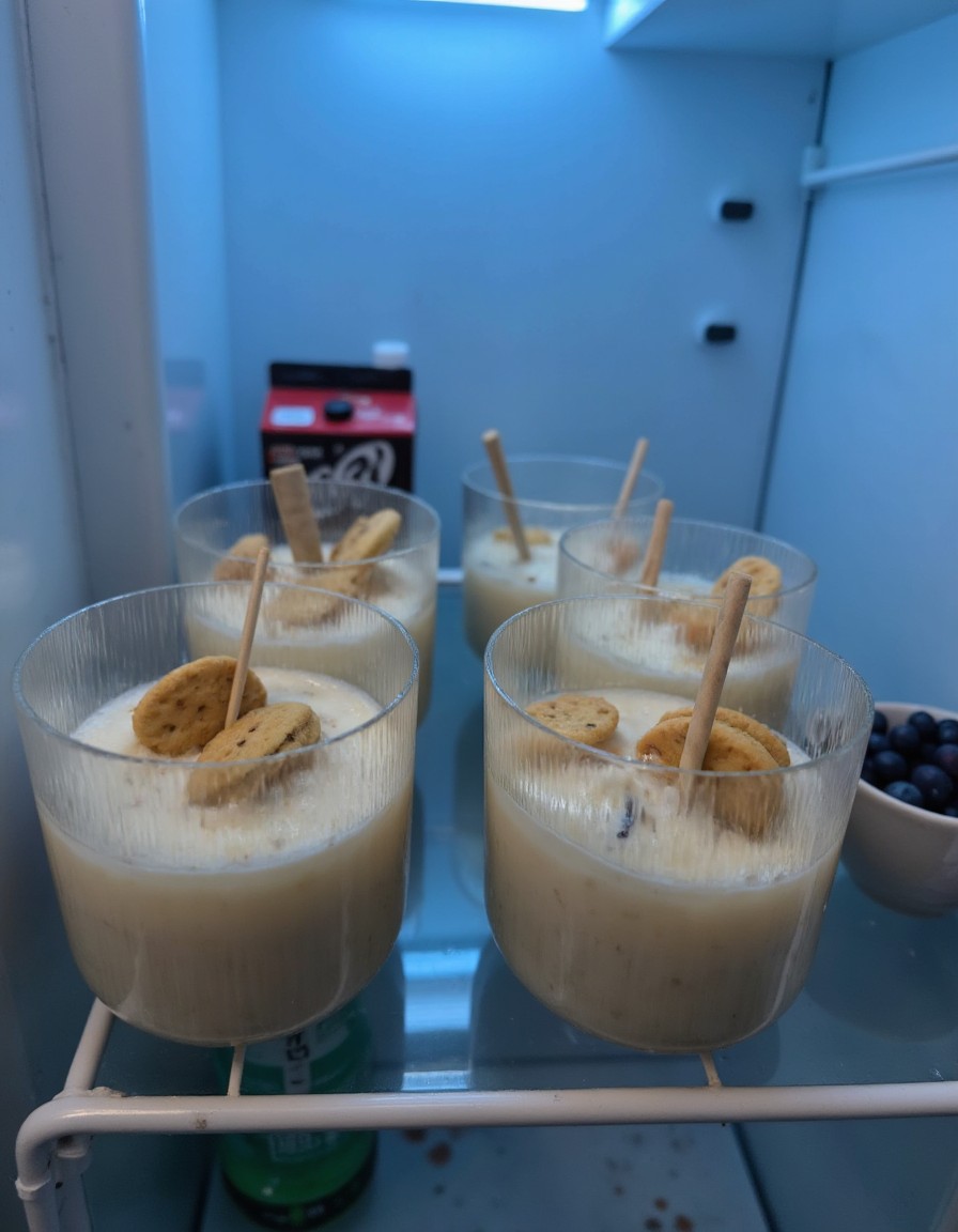 Vier mit Frischhaltefolie abgedeckte Dessertgläser mit Joghurt und Biscoff-Keksen auf einem Kühlschrankregal